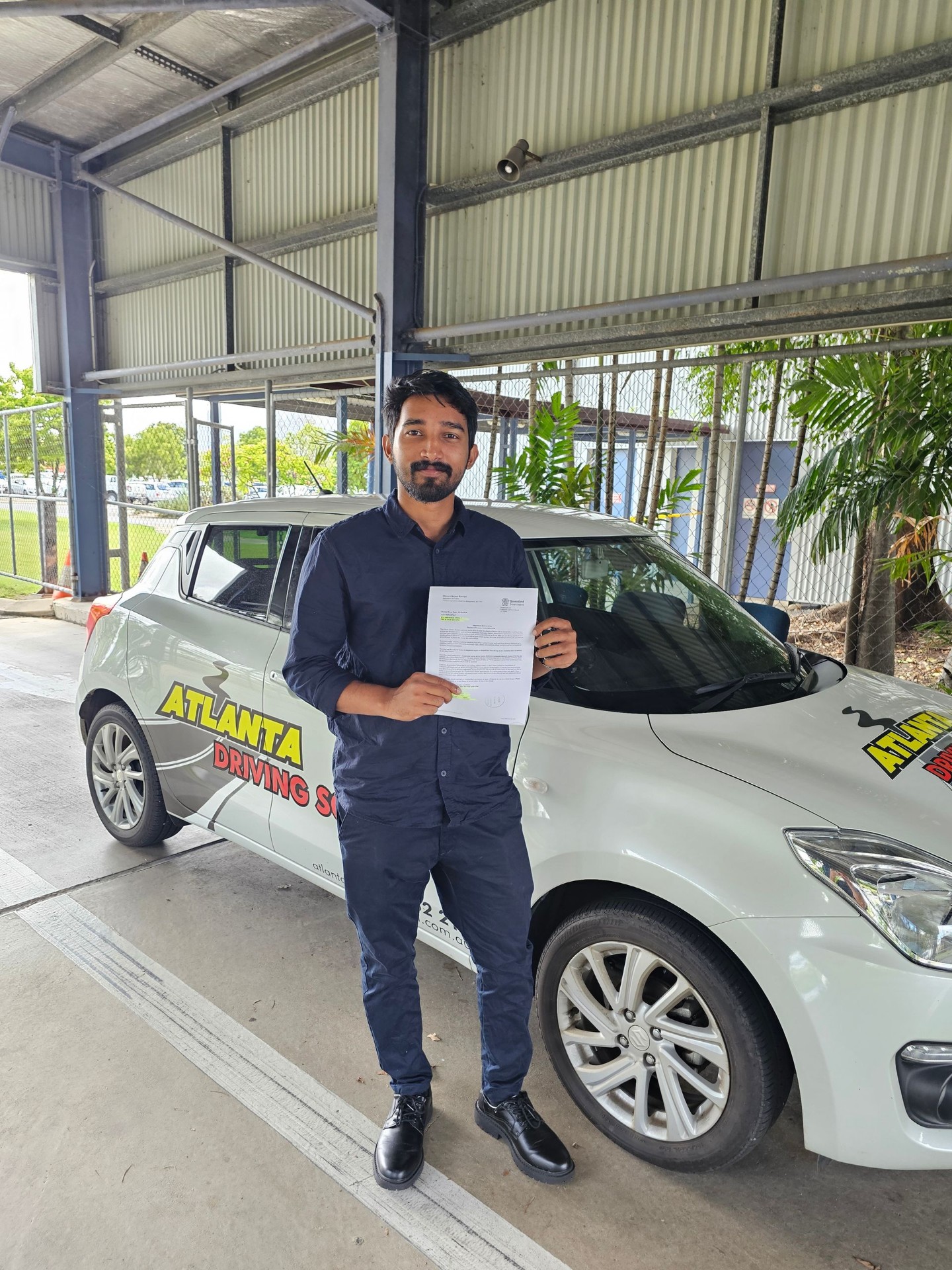 Atlanta Driving School student smiling beside the branded training car at the Rockhampton testing centre, holding his Queensland driver licence receipt after passing the practical driving test